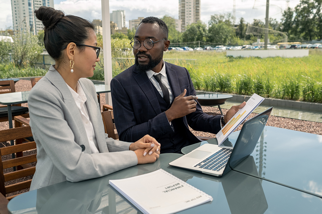 Two young multiracial economists discussing financial paper by table during outdoor meeting